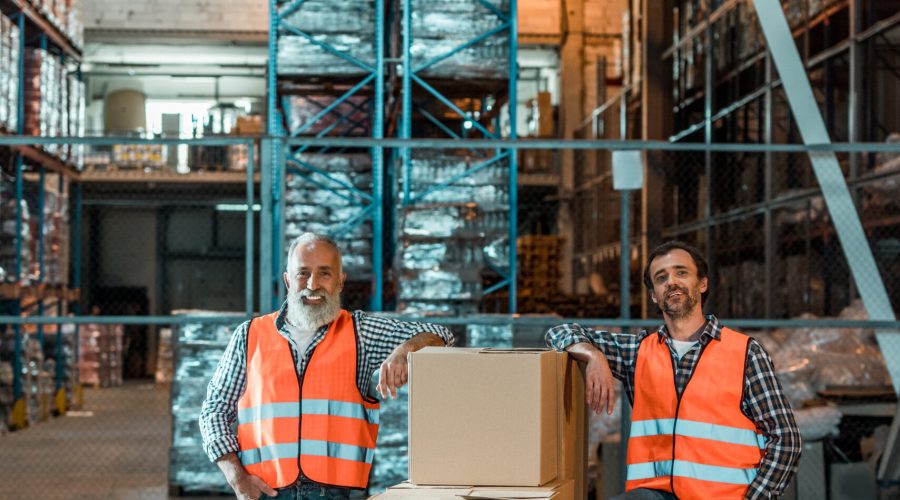 male warehouse workers in vests standing with boxes and smiling at camera