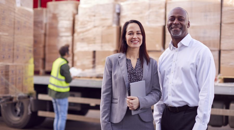 Portrait Of Multi-Cultural Freight Haulage Team Standing By Truck Being Loaded By Fork Lift